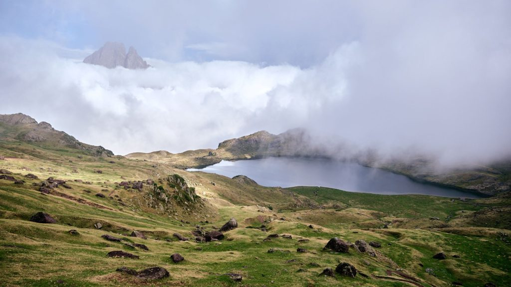 A high alpine tarn surrounded by tussock grassland below Mount Kenya's rocky ridges, with low clouds drifting over the moorland.