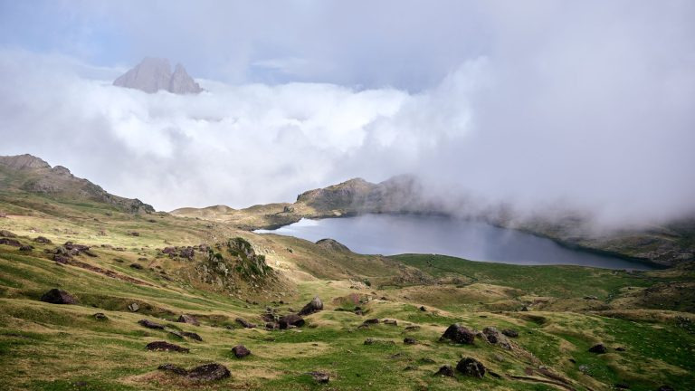 A high alpine tarn surrounded by tussock grassland below Mount Kenya's rocky ridges, with low clouds drifting over the moorland.