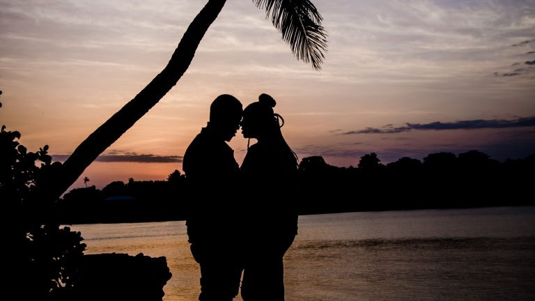 A couple's silhouette stands beneath swaying palm trees as the sun sets over the ocean in Mombasa, Kenya.
