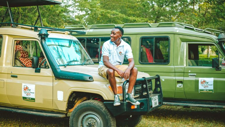 A smiling man sits atop an open safari vehicle as a giraffe grazes nearby on the savannah.