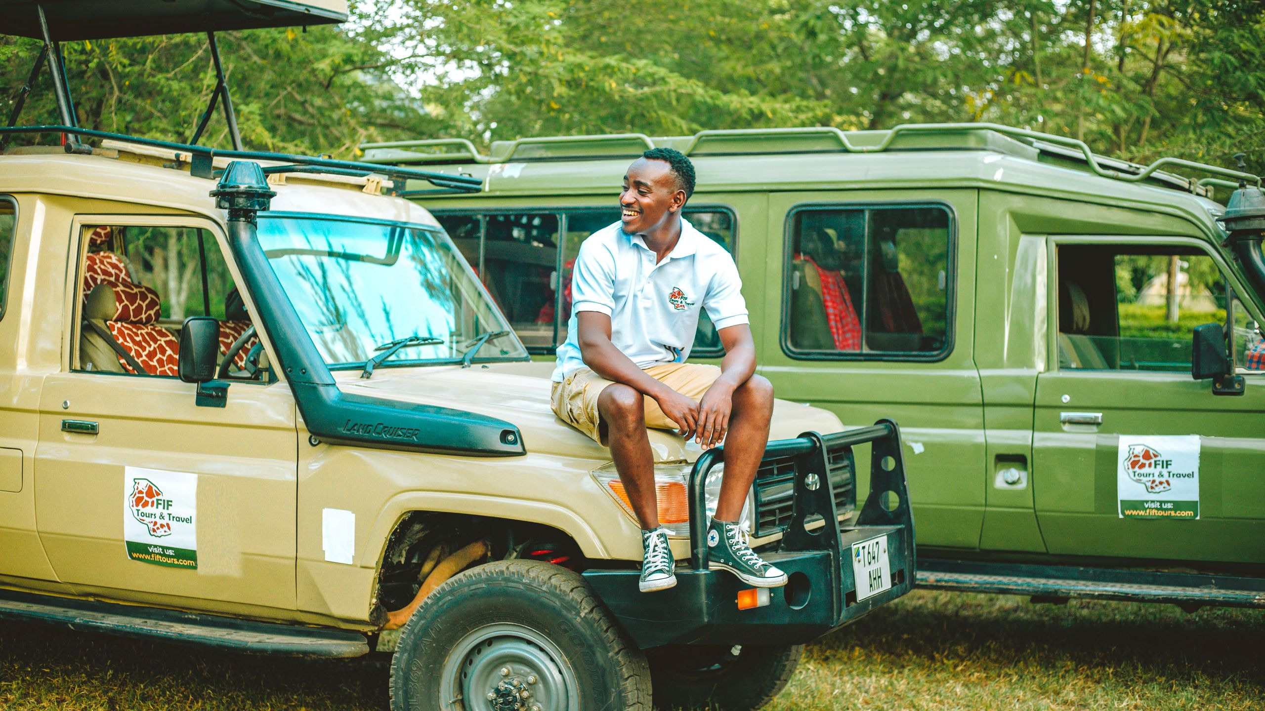 A smiling man sits atop an open safari vehicle as a giraffe grazes nearby on the savannah.