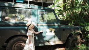 A young girl in a sun hat stands beside a vintage safari jeep in a lush jungle setting