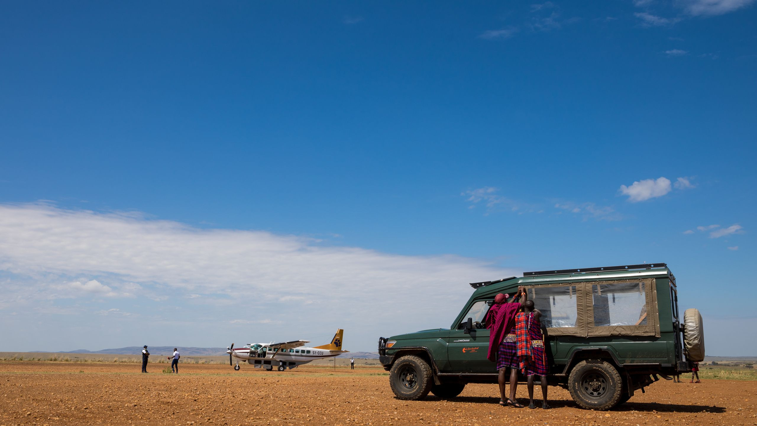 A small bush plane and waiting travelers sit at a remote dirt airstrip in Kenya under a clear blue sky.